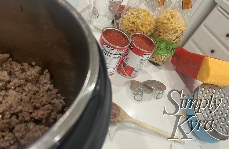 Image shows the cooked meat in the InstaPot with the spoon, grated cheddar, open cans of tomato sauce, and open bags of pasta behind and to the right of it.