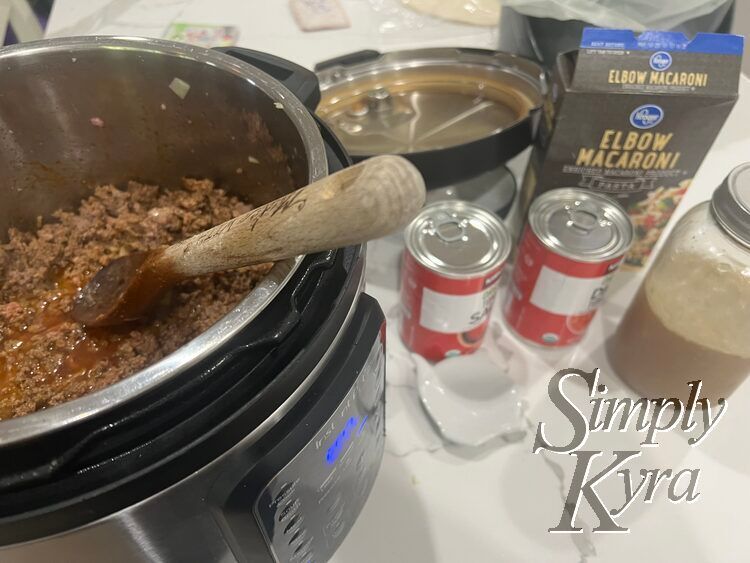 Image shows the InstaPot with cooked and seasoned ground beef in a bit of liquid. The wooden spoon rests in the pot with the rest os the chicken broth, canned tomatoes, elbow macaroni, and the lid blurred in the background.