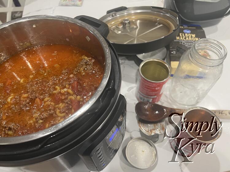 Image is taken from above with an open InstaPot showing cooked ground meat, elbow macaroni, diced tomatoes, and rich chicken broth. To the side lays the wooden spoon, the lid, and the emptied cans and boxes.