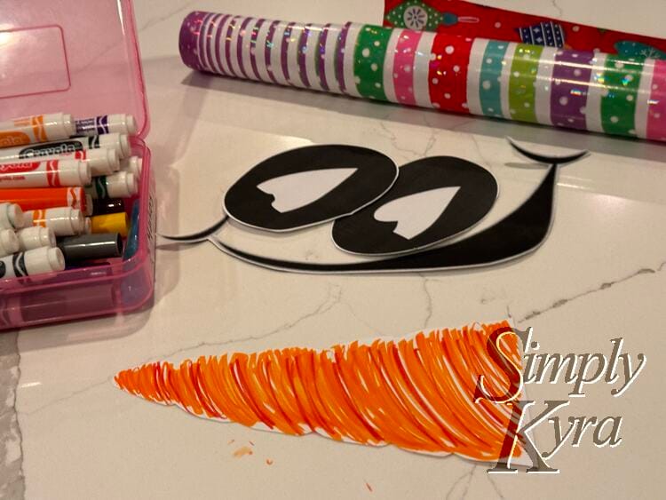 Image shows the cut out eyes and mouth, the wrapping paper, markers, and the colored orange paper carrot.