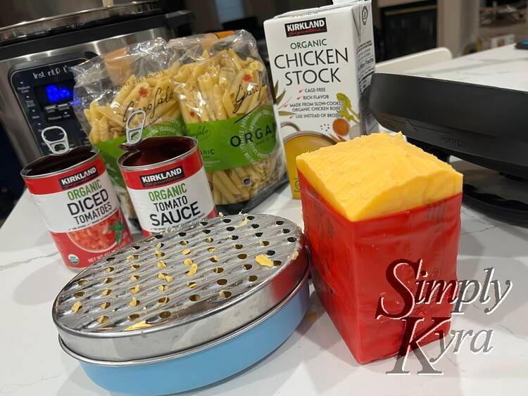 Image shows the pressure cooker in the background with the opened canned tomatoes, pasta, and chicken broth sits in the mid area. In the foreground is a grater filled with sharp cheddar cheese. 