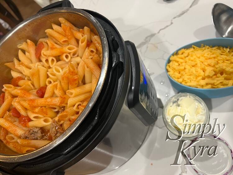 Image shows tomato, meat, and pasta in the pressure cooker with a container of cream cheese, grated cheddar cheese, and a clean spoon off to the side.