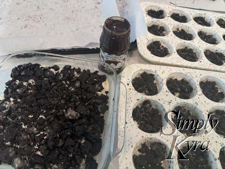 There are two white speckled silicon cupcake trays next to a square glass casserole dish and open jarred of smuckers hot fudge sauce. All have cookies with white ice cream showing through.