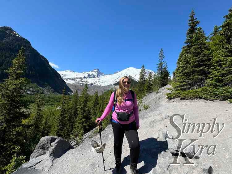 View of me looking at the camera with my hat and hiking pole on gravel with the mountain and glacier behind and trees in the mid. 