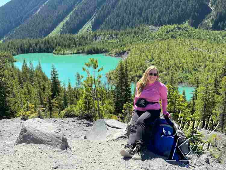 Me sitting posed next the backpack in front of a lake with trees and sunbeam in front and mountains in the background.