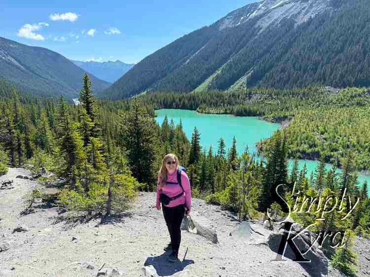 Me standing in front of a lake with trees and sunbeam in front and mountains in the background.