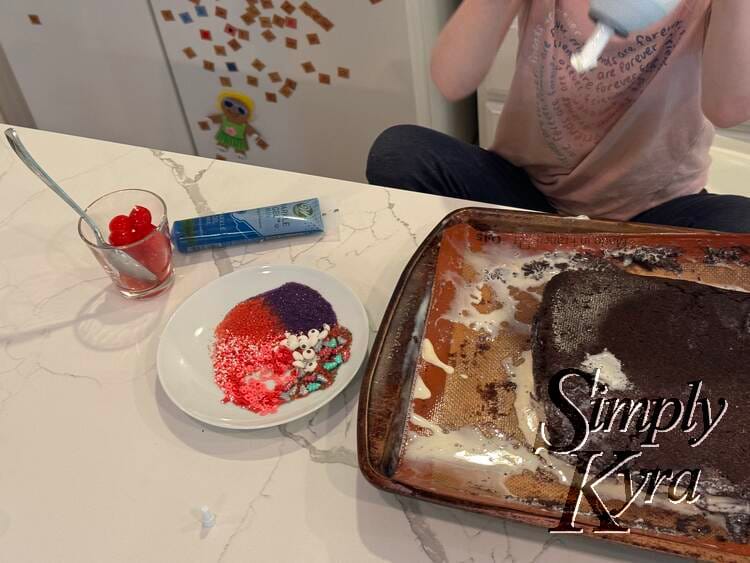 The messy melting but now brownie covered cake is centered on the  silpat lined cookie sheet with whipped cream above and laid on icing writer, cherries, and sprinkles of Zoey's choosing on the left. 