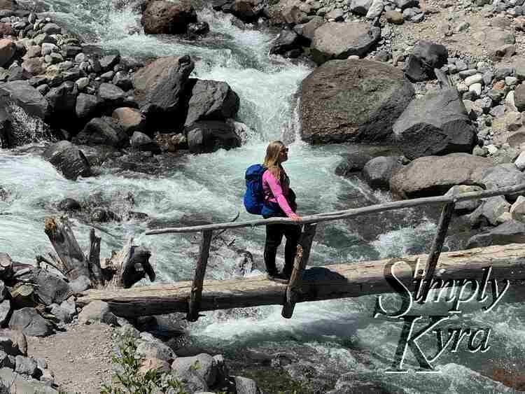 Me standing on skinny bridge with hand on single guardrail over the raging river looking into the distance. 