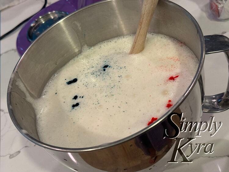 Image shows a silver stand mixer bowl with bubbly white contents and drops of blue and red. 