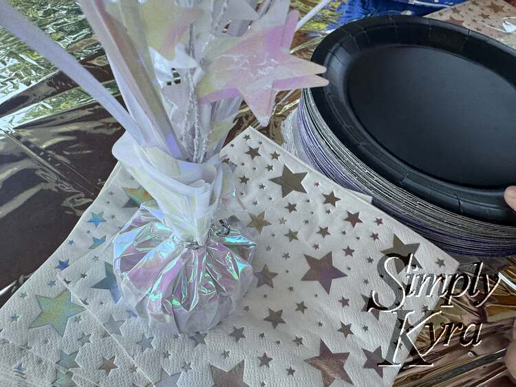 A white silvery and starred weight sits on white napkins bejeweled with silver stars. It sits on a silver tablecloth and next to black plates.