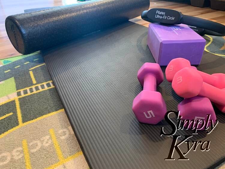 Image shows a pilates mat on a kids car mat with a foam roller, yoga block, magic circle, and two sizes of hand weights laid out on it. 